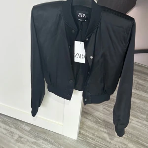 Svart bomberjacka från Zara - Snygg svart bomberjacka från Zara med klassisk ribbad krage, muddar och knappstängning framtill. Jackan har en croppad passform och är tillverkad i ett slätt, lätt glansigt material som ger en clean look. Perfekt för dig som gillar stilrena och trendiga plagg.