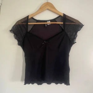 Svart mesh topp med volangärm - Supersöt svart mesh topp från H&M med transparenta volangärmar och liten rosett framtill. Toppen har v-ringning, vågig kant nertill och är gjord i tunn, stretchig syntet. Aldrig använd