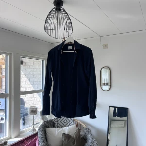 Mörkblå skjorta från Burberry - Snygg mörkblå skjorta från Burberry med klassisk krage och diskret broderad logga på bröstet. Insidan av kragen har det ikoniska rutmönstret i beige och rött. Skjortan är långärmad och tillverkad i mjuk bomull. Perfekt för dig som gillar stilrena plagg med lyxig känsla. Köpt i london!