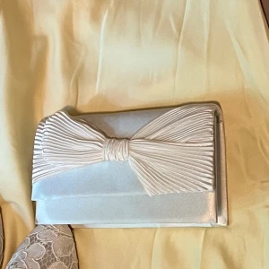 Vit kuvertväska med stor rosett - Snygg vit kuvertväska i satin med en stor, veckad rosett framtill. Perfekt accessoar när du vill sticka ut och addera lite extra elegans till din outfit. Stilren och enkel att matcha med olika looks. Använd vid 1 tillfälle. Nypris 300kr