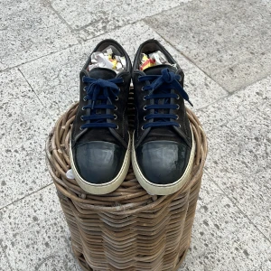 Svarta sneakers från Lanvin i mocka - Snygga svarta sneakers från Lanvin med ovandel i mocka och lackad tå. Skorna har mörkblå snörning och vit sula som ger en clean kontrast. Perfekta för dig som gillar stilrena och exklusiva sneakers med en twist.