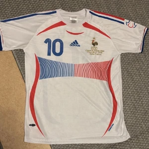 Frankrike VM 2006 fotbollströja - Säljer en vit Frankrike t shirt från Adidas med blå och röda detaljer. Tröjan har korta ärmar och nummer 10 på framsidan samt namnet 'Zidane' på baksidan. Från final 2006 Italien vs Frankrike. Väldigt sällsynt T-shirt!!