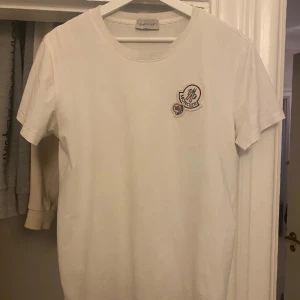 Moncler t shirt - Vit t-shirt från Moncler med klassisk rund hals och korta ärmar. På bröstet sitter två broderade Moncler-loggor i vitt, svart och rött som ger en clean men exklusiv vibe. Tillverkad i mjuk bomull som känns skön mot huden. Perfekt för dig som gillar stilrena märkesplagg.