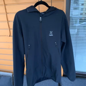 Svart vindjacka från Haglöfs med huva - Svart vindjacka från Haglöfs med minimalistisk design, hel dragkedja framtill och två dragkedjeförsedda fickor. Jackan har en smidig huva och är tillverkad i ett lätt syntetmaterial, perfekt för blåsiga dagar. Snygg logga på bröstet och långärmad modell. Jackan är perfekt för sena sommarkvällar, våren och hösten! Priset kan diskuteras!