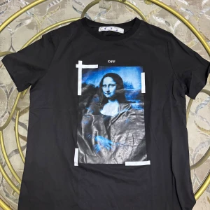 Svart Off-White t-shirt med tryck - Svart t-shirt från Off-White med stort grafiskt tryck i blått och vitt framtill och ikoniska vita pilar på ryggen. Klassisk rund hals och korta ärmar. Materialet känns mjukt och skönt, perfekt för en streetwear-look.