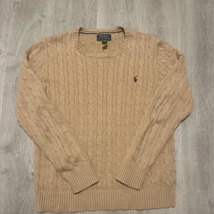 Beige kabelstickad tröja Polo Ralph Lauren - Kabelstickad beige tröja från Polo Ralph Lauren i 100% bomull. Klassisk rund halsringning, ribbade muddar och nederkant samt den ikoniska broderade loggan på bröstet. Perfekt för lager-på-lager och snygg till jeans eller chinos.