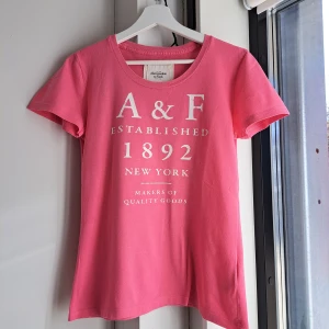 Rosa t-shirt Abercrombie & Fitch - Snygg rosa tröja från Abercrombie & Fitch med vit tryckt text framtill. Klassisk rund halsringning och korta ärmar. Tillverkad i 100% bomull för en skön och mjuk känsla. Fint begagnat skick, ett pyttelitet hål syns på sista bilden. 