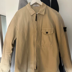 Beige overshirt från Stone Island - Säljer en beige overshirt från Stone Island med klassisk krage, bröstficka med knapp och dragkedja framtill. Mycket sparsamt använd. Färgen är mer mörkbeige än ljus. Storlek Large.
