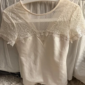 Vit blus med spetsdetaljer - Superfin vit blus med korta ärmar och vackra spetsdetaljer över axlar och bröst. Blusen har rund halsringning och ett elegant, feminint uttryck med genombruten spets upptill. Perfekt för dig som gillar romantisk stil och vill sticka ut lite extra.