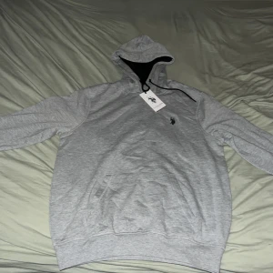 Grå hoodie från U.S. Polo Assn. - Snygg grå hoodie från U.S. Polo Assn. med svart broderad logga på bröstet. Tröjan har huva med svarta snören och en stor magficka. Tillverkad i mjuk bomull, perfekt för chill dagar. Klassisk passform och långärmad modell.