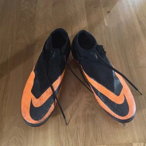 Nike orange hypervenom 46 - Säljer ett par Nike fotbollsskor i orange och svart med stora Swoosh-loggor och snörning. Skorna har en strumpliknande passform och dobbar för gräsplan. Ovandelen är i syntetmaterial och mesh för bra andningsförmåga. Perfekta för dig som vill sticka ut på planen. Storlek 46!