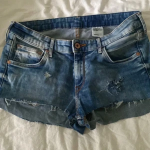 Blå slitna jeansshorts - Snygga blå jeansshorts med slitna detaljer och råa kanter. Klassisk femficksmodell med knapp och dragkedja framtill. Perfekta för varma dagar när du vill ha en avslappnad och trendig look. Lågmidjade