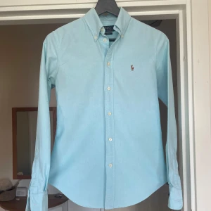 Ljusblå skjorta från Polo Ralph Lauren XS - Snygg ljusblå skjorta från Polo Ralph Lauren i custom fit. Klassisk button-down krage, broderad logga på bröstet och vita knappar. Skjortan är långärmad och tillverkad i mjuk bomull, perfekt för en clean och stilren look. OBS, finns en fläck på höger arm, men går bort i tvätten! Skjortan passar en som är mellan 160 cm till 170 cm, hör av er vid intresse! 