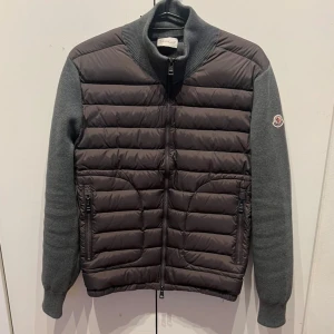 Moncler Cardigan - Snygg pufferjacka från Moncler i mörkgrått med quiltad front och rygg samt stickade ärmar och krage. Jackan har dragkedja framtill, två fickor med dragkedja och Moncler-logga på ärmen. Perfekt för dig som vill ha en stilren och sportig look.
