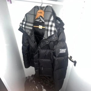 Svart pufferjacka från Burberry - Säljer en svart pufferjacka från Burberry med ärmar som kan tas av och göras till vest. Jackan har en klassisk pufferdesign, dragkedja och hög krage. Insidan har ett rutigt foder i grått och svart. Perfekt för kalla dagar när du vill vara både varm och stilren. Pris kan diskuteras samt möts upp eller fraktar!