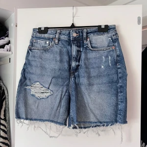 Blå slitna jeansshorts - Snygga blå jeansshorts från H&M med hög midja, slitningar och hål på ena benet samt fransig rå kant nertill. Klassisk femficksmodell i denim med dragkedja och knapp. Perfekta för en avslappnad sommarstil. Önskar du veta exakta mått? Tveka inte att höra av dig!