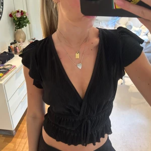 Svart topp med volangdetaljer - Svart topp från Zara med djup v-ringning och korta ärmar . Toppen är croppad med smock och knyt i midjan samt volangkant nertill. Materialet är lätt plisserat. Är mindre i storlek än normal M