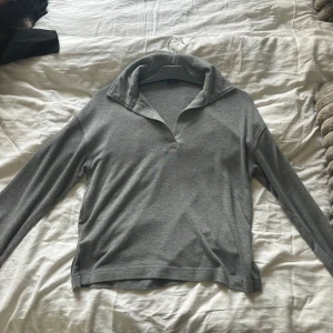 Grå långärmad tröja  - Snygg grå långärmad med avslappnad krage och v-ringad öppning framtill. Tröjan har en enkel och stilren design som passar perfekt till jeans eller kjol. Mjuk och skön, perfekt för lager-på-lager under kyliga dagar.