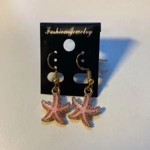 Unika, Handgjorda Rosa sjöstjärneörhängen i guld *Endast 1 Kvar* - Supersöta örhängen med hängande sjöstjärnor i ljusrosa och guldfärgade detaljer. Perfekt för dig som vill ha en lekfull och strandig vibe på din look. Krokarna är guldfärgade och sjöstjärnorna har en glansig finish som verkligen poppar.