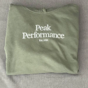Olivgrön Peak Performance sweatshirt - Säljer en olivgrön sweatshirt från Peak Performance med vit broderad logga på bröstet. Tröjan är i mjuk bomull och har en klassisk rund hals. Perfekt för dig som gillar stilrena och sportiga plagg. Endast 149kr