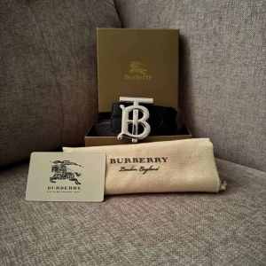 Svart Burberry bälte med silverdetalj - Snyggt svart bälte från Burberry med en stor, ikonisk TB-logga i silver som spänne. Bältet är gjort i skinn och levereras med originalbox, dustbag och certifikat. Perfekt accessoar för att lyfta din outfit med en lyxig touch.