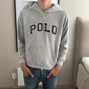 Grå hoodie från Polo Ralph Lauren - Snygg grå hoodie från Polo Ralph Lauren med stor svart POLO-text framtill. Klassisk känguruficka och huva, perfekt för chill dagar. Mjuk bomullsblandning och normal passform. Enkel att matcha med jeans eller joggers.