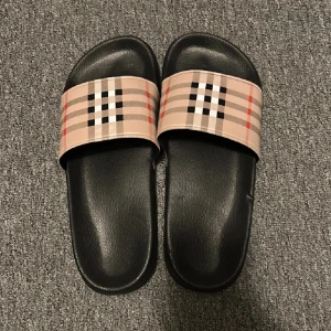 Burberry sandaler med klassiskt rutmönster - Snygga sandaler från Burberry med det ikoniska beige, svarta och röda rutmönstret på remmen. Svart platt sula i syntetmaterial och öppen tå. Perfekta för sommaren och chill dagar. 