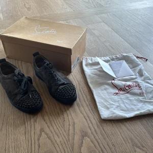 Christian Louboutin Louis Junior Spikes - Snygga mörkgrå sneakers från Christian Louboutin i mocka med svarta nitar på tån. Skorna har rund tå, snörning och platt sula. Kommer med originalkartong och dustbag. Perfekta för dig som vill sticka ut med en edgy och lyxig stil.