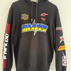 Diesel Alpinestars Hoodie - Svart hoodie från Diesel Alpinestars i storlek M med färgglada racinginspirerade tryck på bröst, rygg och ärmar. Tröjan har huva med snörning och ribbade muddar. 