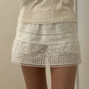 Vit kort kjol med broderade detaljer - Superfin vit kort kjol med broderade hålmönster och volang. Kjolen har en feminin och somrig vibe, perfekt till varma dagar. Den är gjord i bomull och har en söt, luftig känsla. Passformen är normal och kjolen har fina detaljer som ger den en unik look.