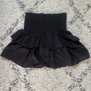 Svart volangkjol från Neo Noir - Supersnygg svart kortkjol från Neo Noir med bred smock i midjan och dubbla volanger. Kjolen har en feminin och trendig look som passar perfekt till sneakers eller boots. Materialet känns lätt och flowy, vilket gör den extra bekväm att bära. Kjolen är också helt oanvänd!