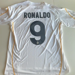 Real Madrid Ronaldo 9 Adidas tröja - Klassisk vit Real Madrid fotbollströja replika med Ronaldo 9 på ryggen. Adidas-logga, klubbmärke och Bwin-sponsor på bröstet. Tunn och luftig ClimaCool-material, kortärmad med gula detaljer längs axlar och sidor. Perfekt för dig som älskar fotboll och vill ha en ikonisk tröja. Endast handtvätt gäller.