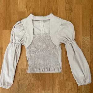 Ljusbeige blus med puffärmar - Supersöt ljusbeige blus med puffiga långärmar och smockad framsida. Toppen har en unik bolero-liknande överdel och hög halsringning. Materialet känns mjukt och har en lätt glans. Perfekt för dig som gillar trendiga detaljer och volym i ärmarna.