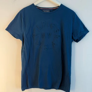 Blå t-shirt Tommy Hilfiger M - Snygg blå t-shirt från Tommy Hilfiger i bomull med rund hals och kort ärm. Framsidan har ett upphöjt tryck med texten 'Hilfiger NY U.S.A.' i samma färg som tyget. Klassisk och stilren modell med diskreta detaljer. Storlek M