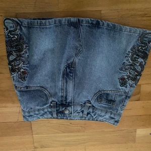 Ed Hardy jeanskjol med drakbrodyr - (Ber om ursäkt för att bilderna är upp och ner vet ej varför det blev så..) Säljer en kort jeanskjol från Ed Hardy med grymma broderade drakar i svart och vitt framtill och stor röd Ed Hardy-logga bak. Kjolen är helt ny från butik helt oanvänd