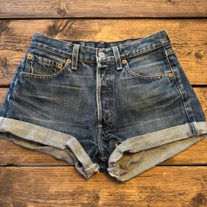Levi's 501 jeansshorts blå denim - Klassiska retro  Levi's 501 jeansshorts i blå denim med upprullade benslut och femficksmodell. Snygg vintagekänsla med slitningar och ikonisk läderpatch bak. Midjestorlek 28 (retro, så skulle säga xs i dagens mått)