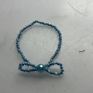 Blått pärlarmband med rosett - Ett sött armband gjort av små blå pärlor med en rosettform framtill och en större pärla i mitten. Perfekt accessoar för att ge din outfit en gullig touch. Armbandet är elastiskt och passar de flesta handleder. Eget gjort!