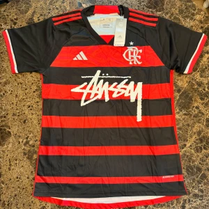 Adidas Flamengo x Stüssy fotbollströja M - Snygg fotbollströja från Adidas i samarbete med Stüssy och Flamengo. Tröjan är svart och röd med breda horisontella ränder, vit Stüssy-logga på bröstet och Flamengo-emblem. Tillverkad i lätt och ventilerande Aeroready-material. Klassisk V-ringning och korta ärmar.