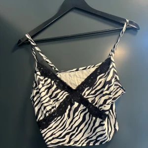 Zebramönstrad linnetopp med spets - Snygg zebramönstrad linnetopp med tunna axelband och svarta spetsdetaljer över bysten. Toppen är vit och svart med croppad passform, perfekt för att sticka ut. Mjuk och lätt i materialet, passar till både jeans och kjol. Stl S/M