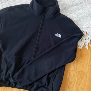 Svart fleece half zip från The North Face - Svart tunn fleece-tröja från The North Face med halv dragkedja och hög krage. Broderad logga på bröstet och praktiska fickor framtill. 