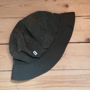 Mörkgrön bucket hat från Wood Wood - Bucket hat i mörkgrönt från Wood Wood med klassisk skärm och ventilerande meshfoder på insidan. Hatten har en liten vit logolapp framtill och är tillverkad i lätt, slitstarkt syntetmaterial. Oanvänd, inköpt på sample sale.