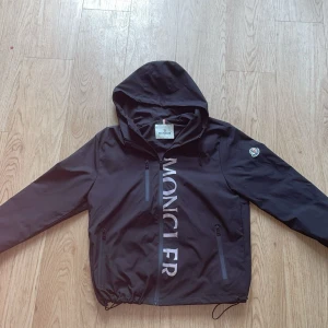 Svart vindjacka från Moncler med huva - Svart vindjacka från Moncler med stor logga framtill, huva och dragkedjor på fickorna. Jackan har justerbar dragsko nertill och klassisk Moncler-patch på ärmen. Perfekt för dig som vill ha en clean och sportig look.
