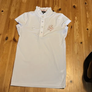 Vit tävlingstopp med pärlknappar från Spooks - Supersöt vit tävlingstopp från Spooks i storlek XS. Tävlingstoppen har korta ärmar, volangkrage och pärlknappar framtill. Detaljer med broderad logga och blommigt tyg på insidan av kragen. Perfekt för sommarens alla tävlingar och träningar. KAN ABSOLUT GÅ NER I PRIS VID SNABB AFFÄR ELLER BUNDLE!