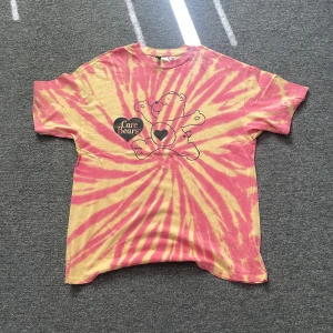 Tie-dye Care Bears t-shirt Divided XS - Säljer en färgglad tie-dye t-shirt från H&M Divided i samarbete med Care Bears. T-shirten är rosa och gul med coolt spiral-mönster och tryck av en Care Bear på bröstet. Skön bomullskvalitet och klassisk passform med korta ärmar.