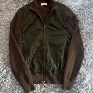 Grön och brun overshirt från Moncler - Snygg overshirt från Moncler i mörkgrönt och brunt. Jackan har stickade ärmar och rygg i ull samt glansig polyester framtill. Dragkedja hela vägen, ståkrage och diskret Moncler-logga på ärmen. Perfekt lager-på-lager-plagg för höst och vår.