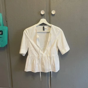 Vit blus med knyt från H&M Divided - Supersöt vit blus från H&M Divided med v-ringning och knytband framtill. Modellen har korta ärmar och volang nedtill som ger en luftig känsla. Perfekt till jeans eller kjol för en fräsch look. Gjord i bomull för en skön och lätt känsla.
