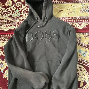 Svart hoodie från Hugo Boss - Svart hoodie från Hugo Boss med stor broderad logga på bröstet. Tröjan har huva med snörning och en klassisk känguruficka framtill. Mjuk insida och ribbade muddar vid ärmslut och nederkant. Perfekt för en chill och stilren look.