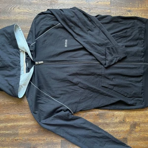 Svart Hugo Boss Zip-up  - Svart zip-up med huva från BOSS med grått foder i huvan och diskreta gråa detaljer längs axlarna. Koftan har dragkedja framtill, två fickor och broderad BOSS-logga på bröstet. Perfekt för en chill och stilren look.