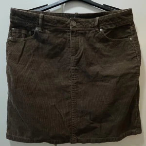 Brun manchesterkjol från Lindex - Snygg brun manchesterkjol från Lindex med klassisk femficksdesign och bälteshällor. Kjolen har jeansinspirerade detaljer med nitar och knapp framtill. Perfekt för dig som gillar retrovibbar och vill ha en cool vardagslook.