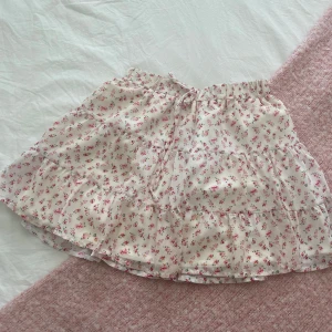 Söt blommig volangkjol med snörning - Supersöt kort kjol med volanger och små rosa blommor på vit botten. Kjolen har snörning i midjan och är perfekt för vår och sommar. Lätt och luftig, passar till sneakers eller sandaler.